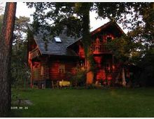 Holzhaus in einem Garten, umgeben von Bäumen. Das Gebäude hat ein Satteldach, einen kleinen Balkon und wird von warmem Sonnenlicht angestrahlt.