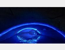 Dunkler Tunnel mit gemauerten Wänden, beleuchtet durch blaue LED-Lichter, die entlang der oberen Rundungen der Bögen angebracht sind. Im Vordergrund ist ein ruhiges Gewässer, das das blaue Licht reflektiert.