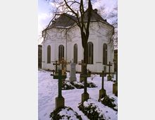 Außenansicht einer weißen Kirche mit hohen, gotischen Fenstern, umgeben von einem verschneiten Friedhof mit mehreren Kreuzen und Grabsteinen. Ein kahler Baum steht prominent vor der Kirche und wirft Äste über das Bild.
