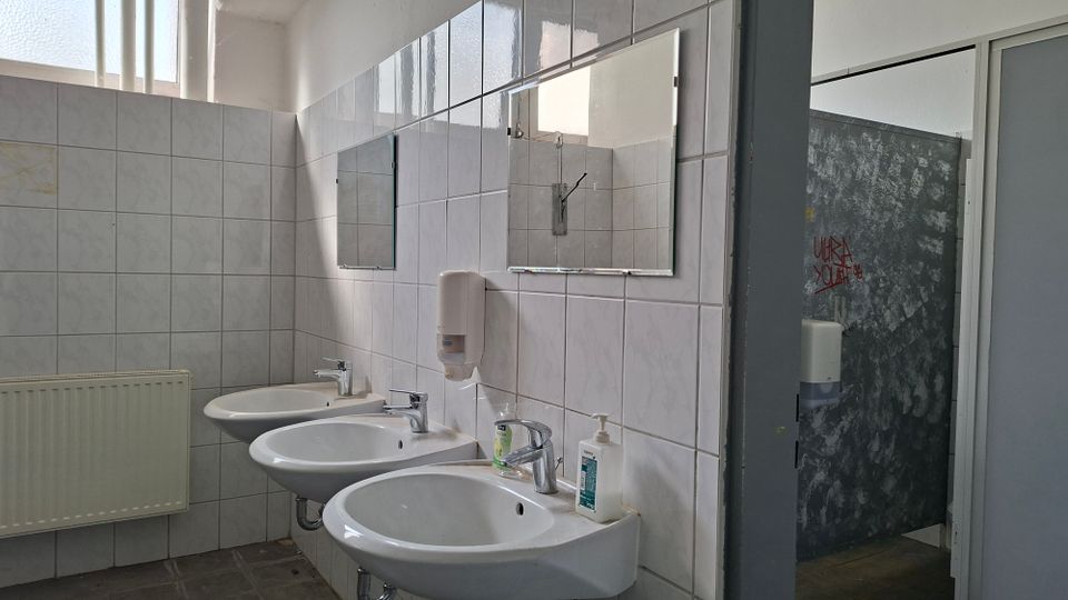 Gefliester Waschraum mit drei wandmontierten Waschbecken, Spiegeln und Seifenspendern, beleuchtet durch ein hohes Fensterband. Rechts ist ein angrenzender Bereich mit grauer Wand und sichtbaren Kabinenabtrennungen zu erkennen.