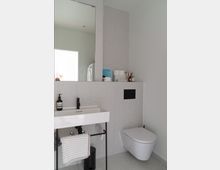 Modernes Badezimmer mit einem freistehenden weißen Waschbecken auf einem schwarzen Metallgestell, einem großen Wandspiegel, einem wandmontierten WC und hellgrauen Fliesen an Wand und Boden. Im Hintergrund sind weitere Badezimmerutensilien und ein beleuchteter Raum sichtbar.