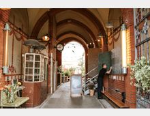 Ein schmaler Durchgang mit Gewölbedecke und roten Backsteinbögen, dekoriert mit Pflanzen, einer Tafel und einer alten Telefonzelle. Im Hintergrund ist ein Außenbereich mit Sitzgelegenheiten sichtbar, ergänzt durch Lichterketten und eine zentrale Uhr.