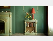 Grünes Wohnzimmer mit antikem Kamin und passender kleiner Bücherkommode. Die Kommode enthält Bücher und dekorative Gegenstände, einschließlich einer Vase mit gelben Blumen und einer rot-orangefarbenen Tischlampe.