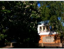 Ein kleines zweistöckiges Haus mit Holzelementen, umgeben von hohen Bäumen. Im Vordergrund steht ein Zaun, und das Haus hat geschlossene Fensterläden sowie ein teilweise sichtbares Dach.