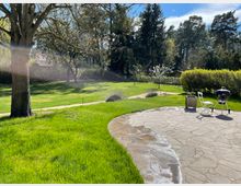 Garten mit gepflasterter Terrasse, darauf ein Grill und zwei Gartenstühle. Im Hintergrund eine große Rasenfläche mit Bäumen, Sträuchern und blühenden Bäumen, umgeben von einer dichten Hecke und Wald am Horizont.