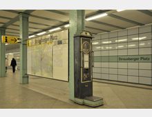 U-Bahn-Station Strausberger Platz in Berlin mit einem historischen Münzwaage-Automaten im Vordergrund, einer gelben Hinweistafel mit Pfeil und einer Übersichtskarte an der Wand. Die Wände sind teils gefliest, teils mit Informationstafeln versehen.