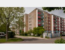 Mehrstöckiges Wohnhaus mit rechteckiger Fassade aus Betonplatten, roten und grünen Balkongeländern. Im Vordergrund ein Gehweg, grüne Bäume, ein Straßenschild mit der Aufschrift „Parsteiner Ring“ sowie ein Stromkasten und ein umzäunter Bereich.