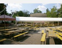 Außenbereich eines Biergartens mit langen Holzbänken und Tischen auf Kiesboden, überdacht durch ein großes weißes Zelt; im Hintergrund sind Bäume, ein Gebäude und ein rot-weiß gestreiftes Sonnensegel sichtbar.