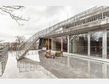Ein modernes Gebäude mit einer großen Terrasse aus Beton, bodentiefen Fenstern und einer Außentreppe aus Metall. Auf der Terrasse stehen Holzmöbel, und im Hintergrund sind Bäume und städtische Gebäude sichtbar.