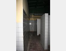 Ein schmaler, gefliester Korridor mit weißen Wandfliesen und braunen Bodenfliesen. An der Decke befinden sich freiliegende Rohre und Kabel, die Wände sind teilweise gelb gestrichen.