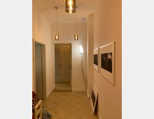 Ein schmaler, modern beleuchteter Flur mit hellen Wänden und beigefarbenem Fliesenboden. An der Decke hängen drei Pendelleuchten mit sichtbaren Glühbirnen, an der rechten Wand sind gerahmte Bilder angebracht, und im Hintergrund ist eine weiße Tür zu sehen.