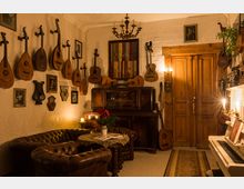Gemütlicher Musikraum mit hölzernen Wänden und einer Sammlung von Saiteninstrumenten, die an den Wänden hängen. Im Vordergrund stehen braune Ledersofas und ein Tisch mit einer Vase, während rechts ein weißes Klavier und links antike Möbel sichtbar sind.