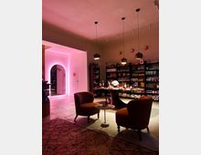 Ein stilvoll eingerichteter Laden mit gedämpfter Beleuchtung, zwei roten Sesseln um einen kleinen Tisch und einem Verkaufsregal im Hintergrund, das mit Lebensmitteln und Getränken bestückt ist. Auf der linken Seite befindet sich ein Durchgang mit pinkfarbenem Licht, das einen modernen Akzent setzt.