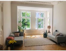 Helles Wohnzimmer mit großen Fenstern, die auf einen grünen Außenbereich blicken. Der Raum verfügt über eine weiße Stuckdecke, einen Holzboden, zwei graue Sofas, Pflanzen, eine Stehlampe und einen strukturierten Teppich.