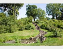 Grüne Parklandschaft mit einer leicht hügeligen Wiese, mehreren Bäumen und Büschen im Hintergrund. Ein kleiner Bachlauf aus Steinen führt zu einem Teich im Vordergrund mit Schilfpflanzen am Ufer.
