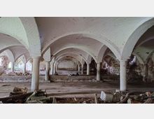 Ehemaliger Gewölbekeller mit weißen Ziegelbögen, gestützt von massiven Säulen. Der Raum zeigt Verfallsspuren, mit herumliegendem Bauschutt und beschädigten Mauern.