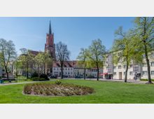 Stadtpark mit Grünflächen, Blumenbeeten und Bäumen im Vordergrund, im Hintergrund eine gotische Kirche mit hohem Turm sowie umliegende historische und moderne Gebäude. Der Himmel ist klar und blau.