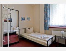 Ein schlicht eingerichtetes Einzelzimmer mit einem Bett, einem runden Nachttisch mit Lampe und einem Kleiderständer. Der Raum hat rote Teppichböden, helle Wände, ein großes Fenster mit blauen Vorhängen und einen Spiegel, der einen Teil des Zimmers reflektiert.