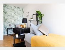 Schlafzimmer mit zwei Einzelbetten, dekoriert mit unterschiedlichen Kissen und Decken in Gelb-, Grau- und Beigetönen. Die grüne Wandtapete mit Blattmuster, schwarze Regalkonstruktionen mit Pflanzen und Accessoires sowie Holzfußboden verleihen dem Raum eine moderne und natürliche Atmosphäre.