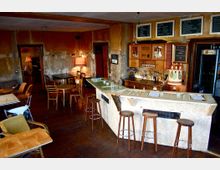 Gemütliche Bar in einem rustikalen Raum mit abgenutzten Wänden, einer zentralen Theke mit Barhockern, Holztischen und -stühlen sowie einer Liste von Getränken auf Tafeln an der Wand. Die Einrichtung wirkt einfach und vermittelt eine entspannte Atmosphäre.