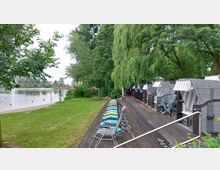 Holzterrasse mit aufgereihten Strandkörben und bunten Liegestühlen neben einer Grünfläche mit Blick auf einen Fluss; im Hintergrund Bäume und Schilfdächer über Sitzplätzen.