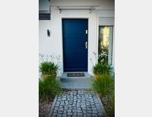 Eingangsbereich eines Wohnhauses mit weiß verputzter Fassade und dunkelblauer Haustür unter einem kleinen Vordach. Ein schmaler Kopfsteinpflasterweg führt zur Stufe vor der Tür, flankiert von bepflanzten Kübeln und Ziergräsern; rechts ist ein Fenster mit Rollladen zu sehen.