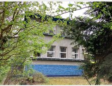 Zweistöckiges Gebäude mit grauer Fassade und einer unteren Wandverkleidung aus blauen Fliesen, umgeben von dichtem Grün und Bäumen im Vordergrund. Die Fenster sind rechteckig und gleichmäßig angeordnet.