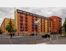 Mehrstöckiges Wohnhaus in warmen Gelbtönen mit roten Balkonen an einer Straßenecke, daneben ein oranger Gebäudeteil mit der Aufschrift „Thoughts Become Things - Happy Hostel“. Im Vordergrund eine Kreuzung mit Straßenschildern, Ampeln und mehreren Fußgängern sowie Autos.