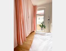 Helles Schlafzimmer mit Holzdielenboden, weißen Bettbezügen und großen, rosafarbenen Vorhängen, die einen Teil des Fensters verdecken. Das geöffnete Fenster mit Rundbogen führt zu einem kleinen Balkon, neben dem eine grüne Zimmerpflanze steht.