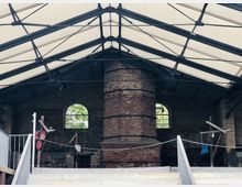 Innenansicht einer historischen Industriehalle mit Backsteinwänden, einem großen gemauerten Schornstein in der Mitte und bogenförmigen Fenstern. Die Decke besteht aus einer Konstruktion aus Stahlträgern und lichtdurchlässigen Paneelen.