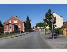 Straßenszene in einer Wohngegend mit einem roten Backsteinhaus links, einem Gehweg mit Straßenlaternen und kleinen Bäumen sowie weiteren Häusern im Hintergrund. Ein Verkehrsschild „Halteverbot“ steht rechts im Bild, und der Himmel ist klar und blau.