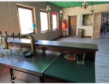 Ein schlicht eingerichteter Gastraum mit einem Tresen im Vordergrund, ausgestattet mit Zapfhähnen und Gläsern. Im Hintergrund stehen mehrere Holzstühle und Kisten mit Getränken; an der Wand hängen Lampen und eine orangefarbene Warnweste.