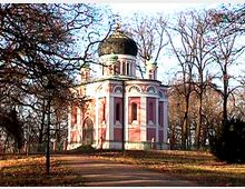 Eine kleine, rosafarbene Pavillon-ähnliche Kapelle mit weißer Verzierung und einer schwarzen Kuppel, umgeben von laublosen Bäumen in einer herbstlichen Parklandschaft. Ein Weg führt zur Kapelle, die im Zentrum des Bildes steht.
