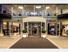 Eingangsbereich eines modernen Gebäudes mit großer Glasfront, einer Drehtür in der Mitte und zwei offenen Schwingtüren seitlich. Beleuchtete Pflanzenkübel stehen links und rechts des Eingangs, im Hintergrund sind Sitzbereiche und eine Wendeltreppe sichtbar.