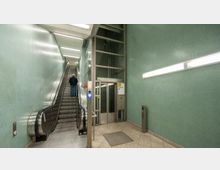 Treppenhaus einer U-Bahn-Station mit einer Rolltreppe auf der linken Seite und einem Aufzug rechts. Wände mit grünen Kacheln, beleuchtet durch helle Neonleuchten.