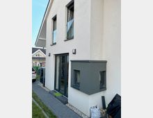 Zweistöckiges modernes Wohnhaus mit heller Putzfassade und grauen Fensterrahmen. Im Vordergrund ist ein kleiner Erker mit Fenster sowie eine gepflasterte Einfahrt und angrenzende Grünfläche sichtbar.