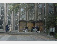 Außenansicht eines modernen Kirchengebäudes mit geometrisch angeordneten, schmalen Fenstern und detaillierten Bronzeflügeltüren, die Reliefs zeigen. Vor dem Eingang stehen mehrere Personen, darunter ältere Menschen mit Rollatoren und eine Person im Rollstuhl, umgeben von herbstlichem Laub.