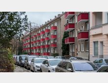 Straßenszene mit mehreren Reihen von Wohngebäuden im klassischen Baustil mit roten Balkonen und beigen Fassaden, flankiert von geparkten Autos auf beiden Seiten der Straße. Im Vordergrund sind Bäume und Gehwege sichtbar, der Himmel ist bewölkt.