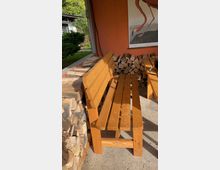 Holzbank unter einem überdachten Bereich, umgeben von gestapeltem Brennholz. Im Hintergrund eine orangefarbene Wand mit einem Bild eines Flamingos, daneben Blick auf einen Garten mit Bäumen.