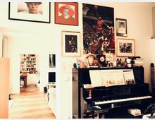 Wohnzimmer mit einer Mischung aus künstlerischer und persönlicher Einrichtung. Im Vordergrund ein schwarzes Klavier mit Notenbüchern darauf, an der Wand darüber Gemälde und Fotografien, im Hintergrund ein Raum mit Bücherregalen und einem Schreibtisch.