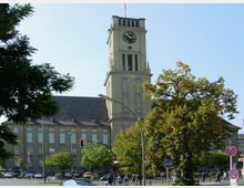 Historisches Rathausgebäude mit Uhrturm im Zentrum des Bildes, umgeben von Bäumen und einer belebten Straße mit Verkehrszeichen, Autos und Passanten. Das Gebäude hat eine Fassade mit hohen Fenstern und ein dunkles Satteldach.
