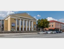 Das Bild zeigt das Friedrich-Wolf-Theater, ein klassizistisches Gebäude mit gelber Fassade, weißen Säulen und großen Fenstern. Vor dem Theater befinden sich Stufen, eine gepflasterte Straße sowie geparkte Autos, mit umliegenden Häusern und einem Baum im Hintergrund.