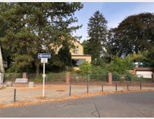 Straßenansicht mit einem Gehweg, auf dem ein Schild mit der Aufschrift „Einbahnstraße“ steht, daneben zwei Parkbänke und Poller. Im Hintergrund ein Wohnhaus mit gelber Fassade, umgeben von Bäumen und dichter Vegetation, eingefasst durch einen niedrigen Zaun aus Metall und Backstein.