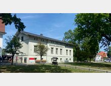 Zweistöckiges Gebäude des Dahmelandmuseums in heller Putzfassade mit mehreren Fenstern und Schrägdach, umgeben von Bäumen und einer Grünfläche. Im Vordergrund stehen Bänke, ein kleiner Spielwagen und ein gepflasterter Weg führt zum Eingang.