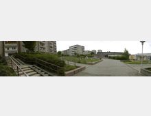 Wohnkomplex mit mehreren mehrstöckigen Plattenbauten und einer zentralen gepflasterten Außenfläche, umgeben von Grünanlagen, Treppen mit Geländern und kleinen Gebäuden. Im Hintergrund befinden sich weitere Wohngebäude und ein Brunnen in einer Rasenfläche.