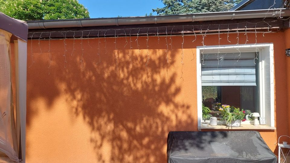 Außenwand eines eingeschossigen Hauses mit orangefarbenem Putz, Dachrinne und einer Lichterkette entlang der Traufe. Rechts ein weiß gerahmtes Fenster mit Fensterbank, auf der Pflanzen und kleine Dekoration stehen; darunter ein abgedeckter Grill, im Hintergrund Baumschatten.