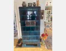 Ein dunkelblau glasierter Kachelofen mit rechteckigen Kachelfeldern und einem metallenen Lüftungsgitter in der Mitte, dekoriert mit kleinen Anhängern. Oben auf dem Ofen stehen Keramikkrüge, daneben befindet sich eine Wandkerze, und rechts davor ein Strauß bunter Blumen in einer Glasvase.