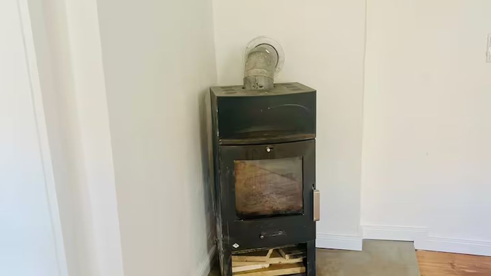 Schwarzer Kaminofen in einer Zimmerecke, auf einer rechteckigen Betonplatte vor weißer Wand. Der Raum hat einen Holzdielenboden und ein kurzes Abzugsrohr führt nach oben in die Wand.