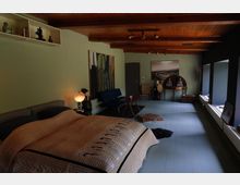 Schlafzimmer mit Holzbalkendecke und grünen Wänden, eingerichtet mit einem Doppelbett, einem blauen Sofa, einem kleinen Beistelltisch und dekorativen Elementen wie Statuen und Bildern. Große Fenster auf der rechten Seite lassen Tageslicht in den Raum.