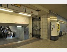 U-Bahn-Station mit Fliesenwänden, einem Fahrkartenautomaten rechts, einer verschlossenen Gittertür und einer Rolltreppe auf der linken Seite. Wände und Automaten sind mit Graffiti bemalt, Sitzbänke und ein Hinweisschild befinden sich im Hintergrund.